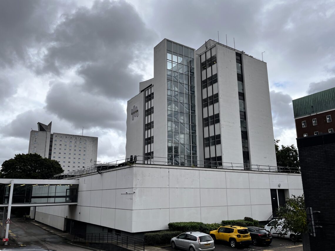 A tall, modern white office building with large glass windows stands under a cloudy grey sky. Three cars, including a yellow one, are parked in front. Another white building and a green-roofed structure are visible in the background. Trees line the scene.