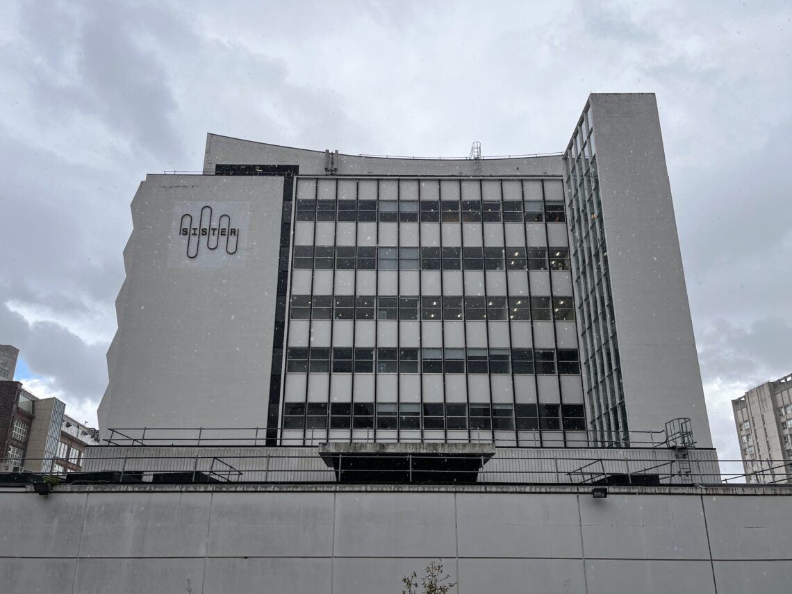 A tall, modernist white building with many windows and a zigzag side façade is seen under a cloudy sky. Snow is lightly falling. The letters “SYSTEM” are displayed vertically on the left side. Other buildings are partially visible on the sides.