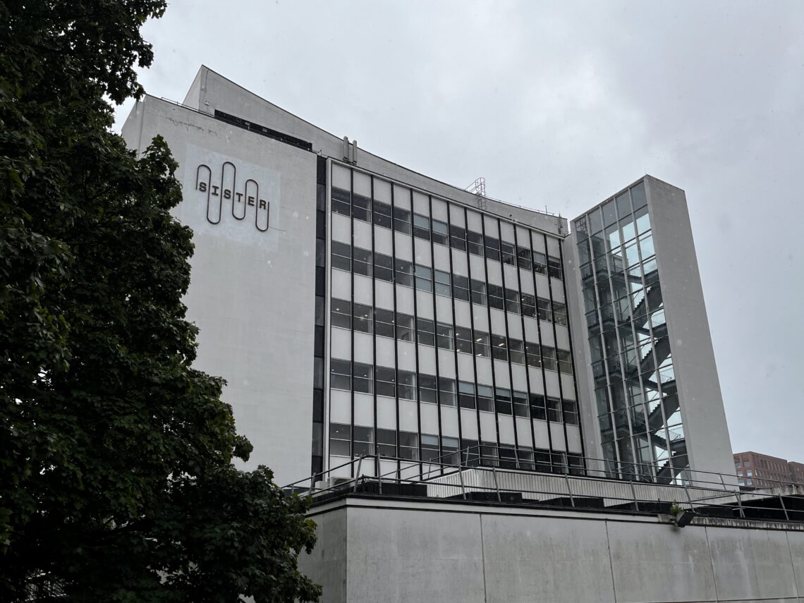 A modern, multi-storey concrete building has large glass windows and a protruding glass stairwell. The word “ASTER” appears in black letters on the upper left corner. A leafy green tree partially obscures the lower left of the structure. The sky is grey.