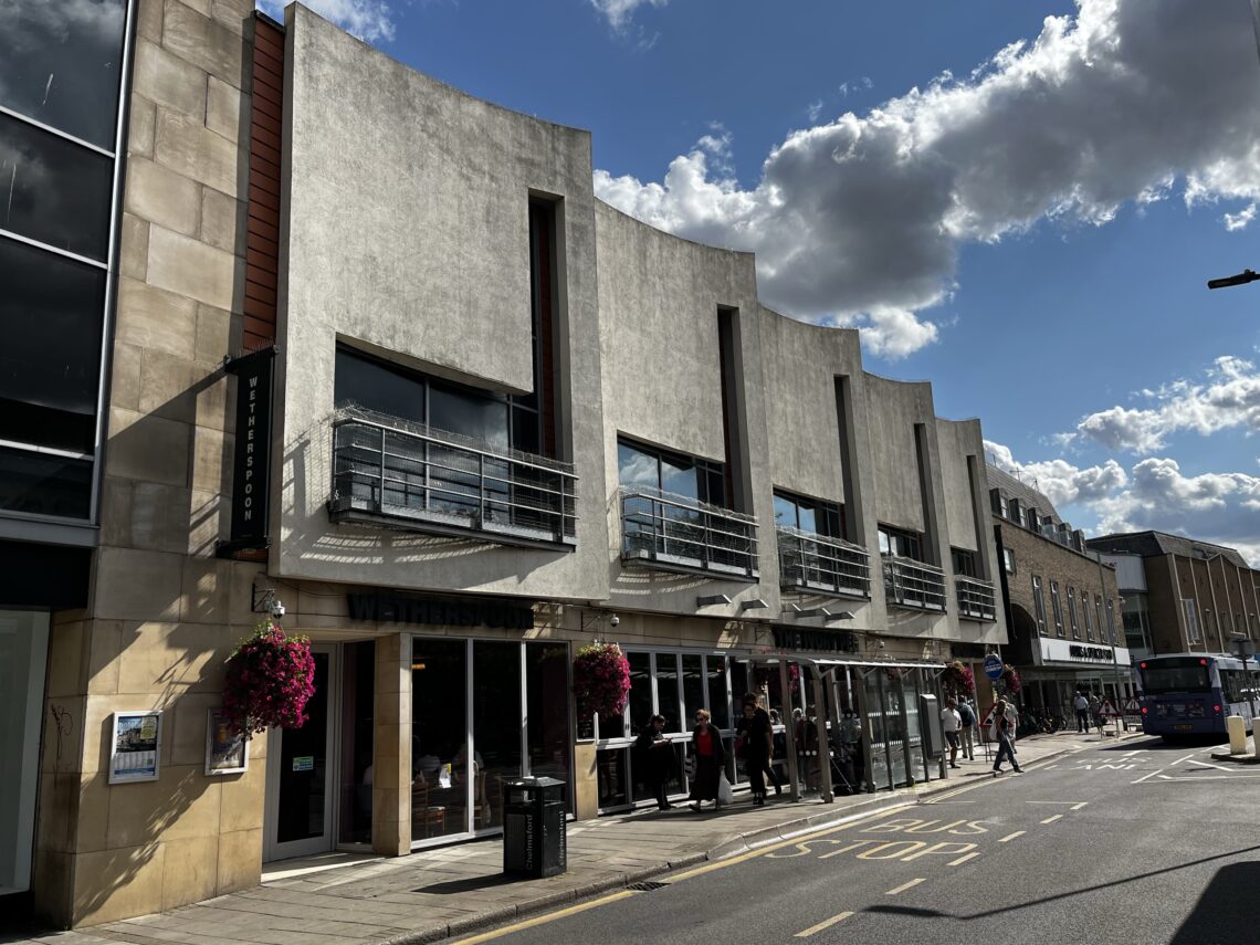 A modern, white, wave-shaped building with large windows and metal railings houses a Waitrose shop. Hanging flower baskets decorate the exterior. People walk outside, and a bus approaches on the sunny street lined with clouds in the blue sky.