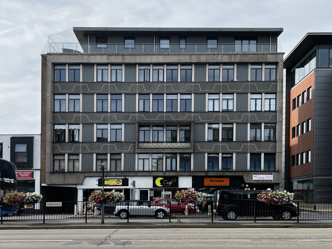 A five-storey modern building with geometric grey facade patterns, large windows, and ground-floor shops. Cars and a van are parked in front. Hanging flower baskets decorate the street, and adjacent buildings frame the scene.
