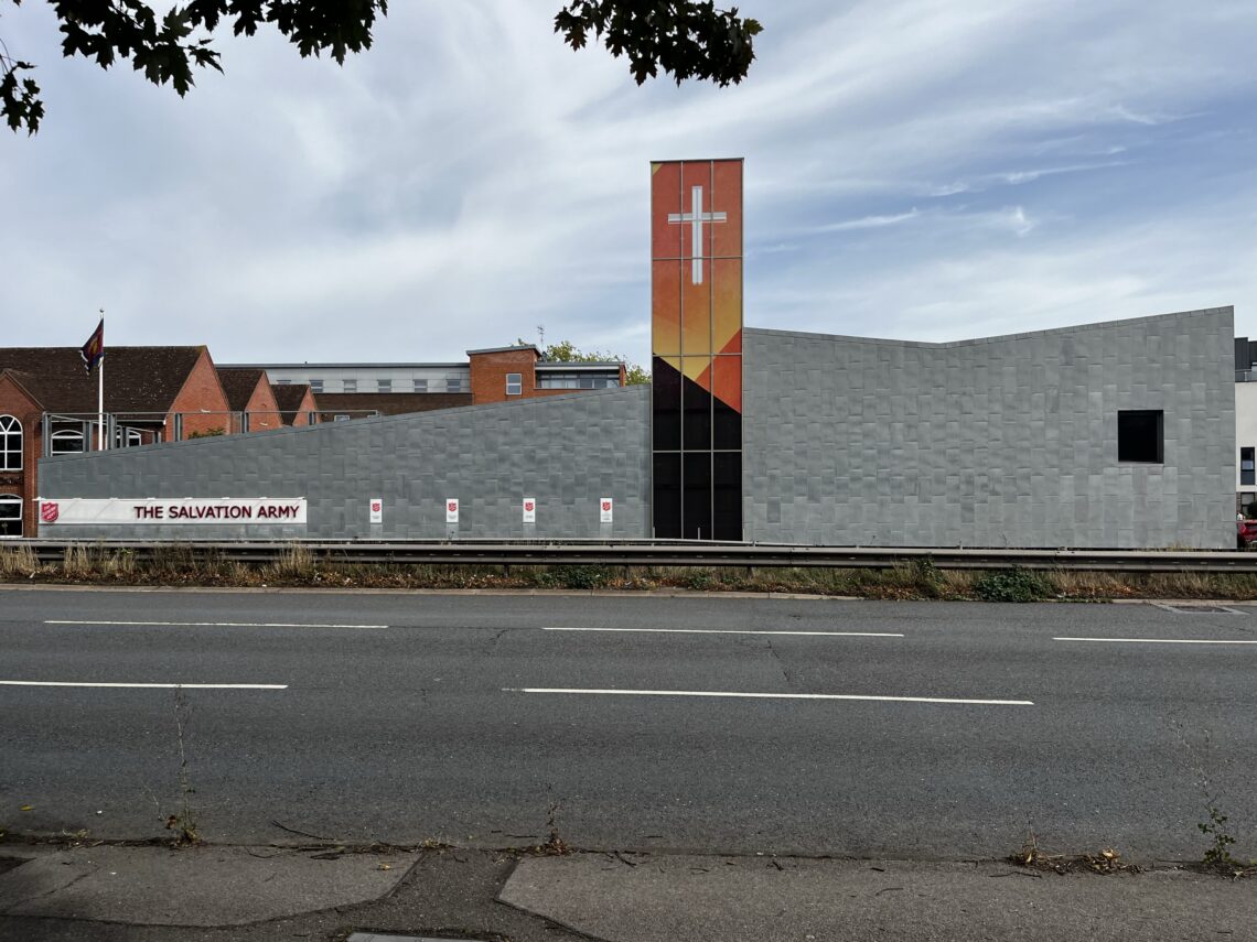 A modern Salvation Army building with a grey, angular façade and a tall tower featuring a white cross on a red and orange gradient. A sign with The Salvation Army is visible along the roadside, with a few trees framing the top edge.