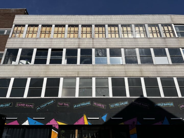 A three-storey building with boarded-up top-floor windows, large white-framed windows on the middle and lower floors, and a colourful geometric mural along the bottom, with painted signs advertising indoor games like escape rooms and shuffleboard.