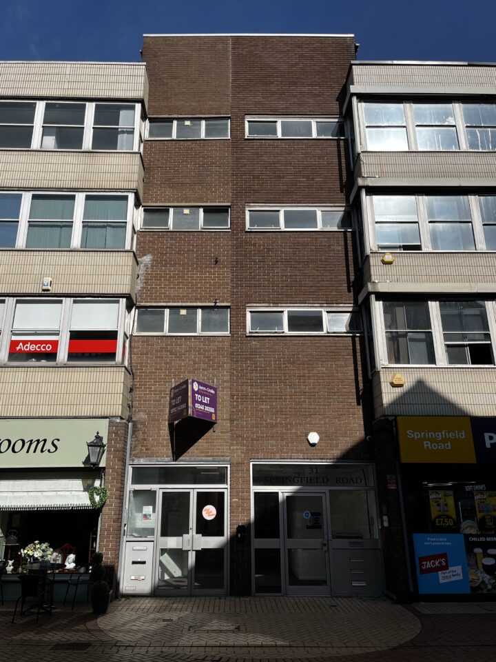 A four-storey brown brick office building with two glass doors at the ground level, flanked by retail shops. A purple “To Let” sign hangs above the entrance. The building has many windows, some reflecting the blue sky.
