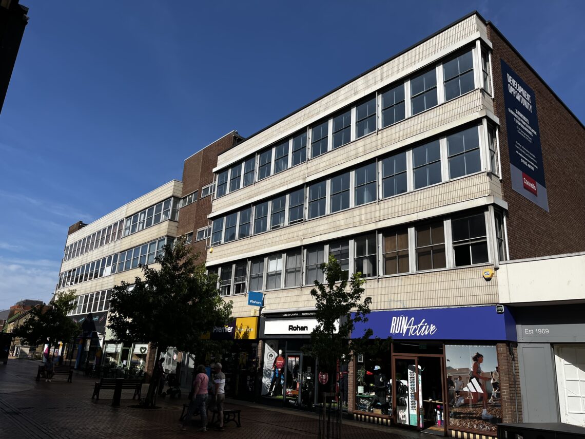 A row of three-storey commercial buildings with shops on the ground floor, including Rohan and RUN Active, under a clear blue sky. People walk along the brick-paved pedestrian street, and leafy trees line the pavement.