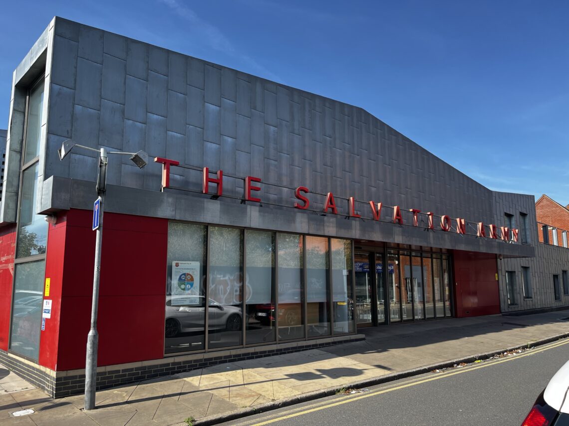 A modern Salvation Army building with a metal facade and red accents. Large windows line the front, and THE SALVATION ARMY is displayed in bold red letters. The building sits on a sunny urban street with a clear blue sky above.