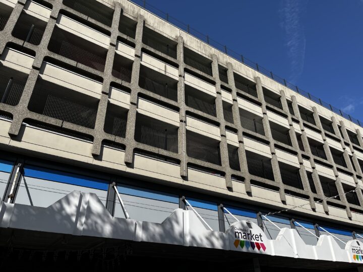 A multi-storey concrete car park with geometric, grid-like openings stands above a street-level market, which has a white awning and colourful market signage. The sky is clear and blue, with sunlight casting shadows on the building façade.