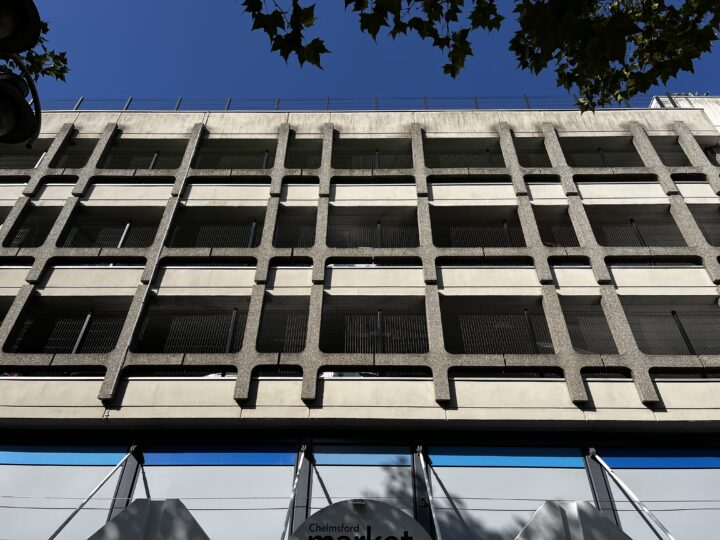 A low-angle view of a concrete building with repetitive square grid-like openings and balconies. The sky above is clear and blue, with leafy tree branches partially visible at the top right corner. Shadows from the trees fall across the building.