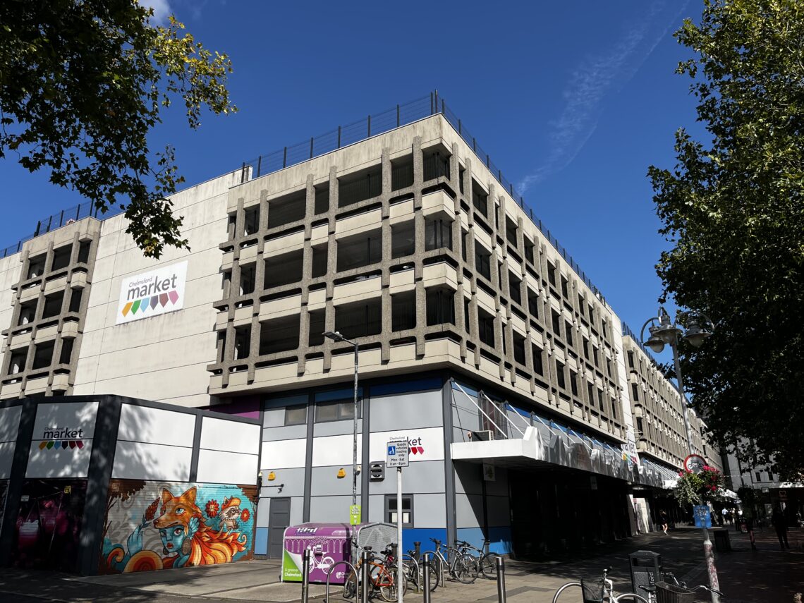 A large, multi-storey concrete car park with a “market” sign on the side. Colourful murals and bikes are near the entrance; trees frame the image. The sky is clear and blue, casting shadows on the building.