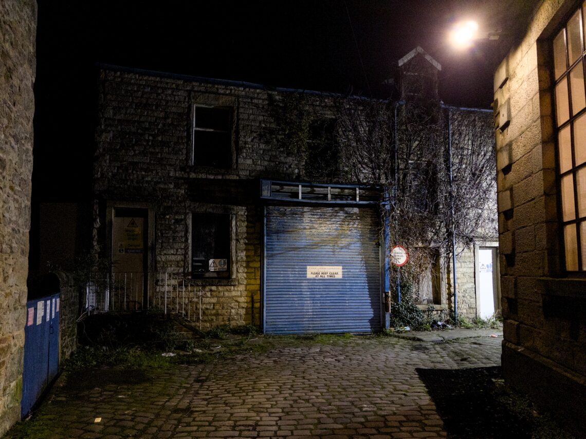 A dimly lit alley at night features an old stone building with boarded-up windows and a blue metal roller shutter. A bright light illuminates part of the building on the right, whilst overgrown plants and uneven cobblestone ground add to the abandoned atmosphere.