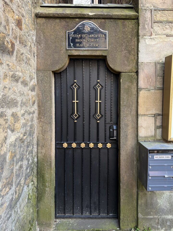 A black, ornate door set in a stone frame with brass decorations and a plaque above reading “SULTAN OF LANCASTER BROCK STREET FLATS 1-4”. A keypad is on the right, and a letterbox unit with labelled boxes is mounted on the stone wall beside the door.