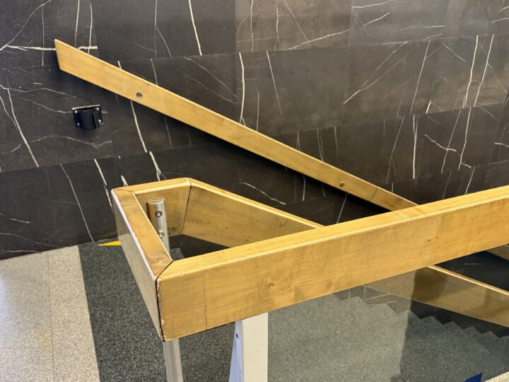 A wooden bannister with an angular corner is shown in the foreground, while another wooden bannister runs diagonally along a dark, marble-patterned wall in the background. The floor is grey with a textured surface, and part of a staircase is visible.