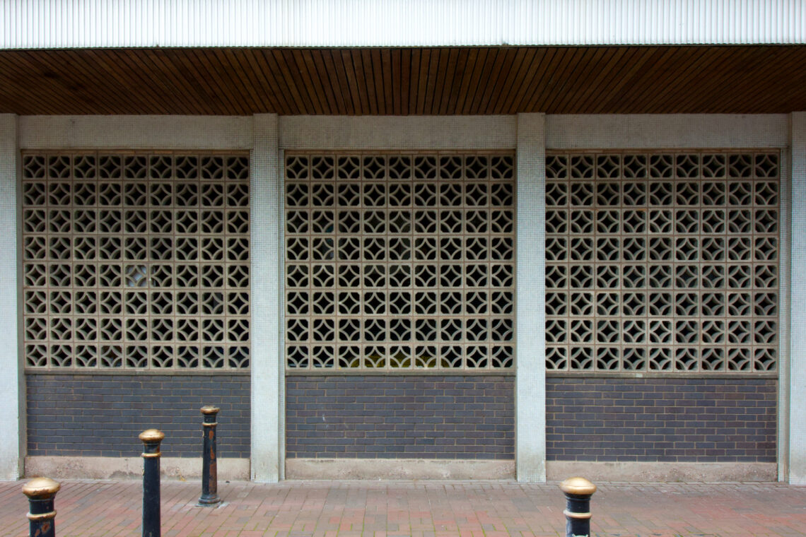 A building façade with three sections of decorative concrete breeze blocks above a base of dark brown bricks. Four black metal bollards with gold tops are in front on a red brick pavement. A metal panel and wooden overhang are above the wall.