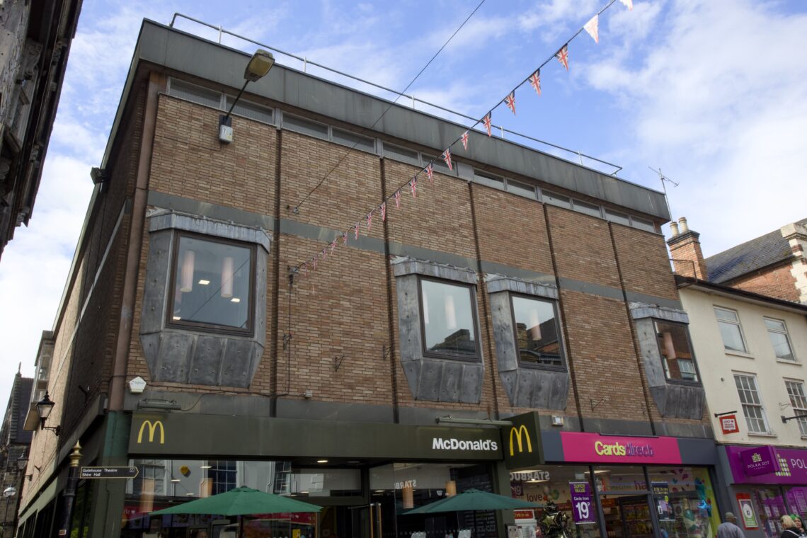 A brick building with three large windows on the upper floor houses a McDonald's and a Card Factory at street level. Small triangular flags hang across the street. Outdoor umbrellas are visible at the bottom of the image.