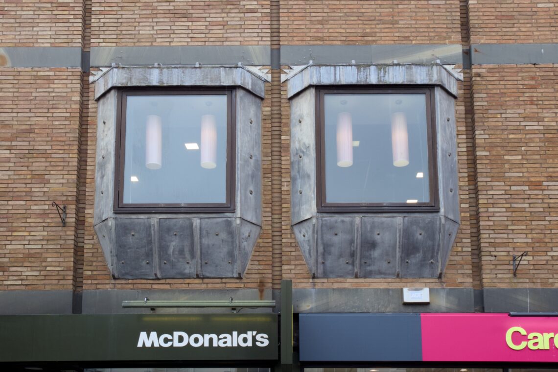 Two large square windows with metal frames are set in a brick building above a McDonald’s sign. Each window shows two cylindrical ceiling lights inside. The bottom border of a pink and yellow shop sign is also partially visible to the right.