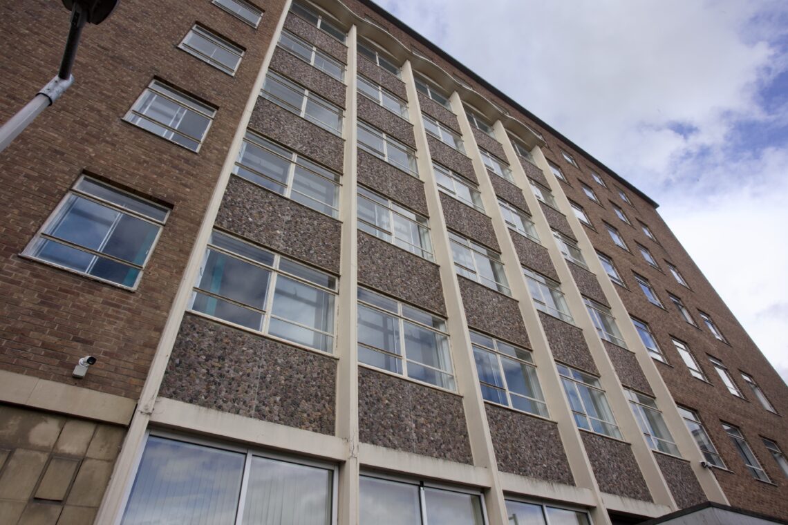 Low-angle view of a multi-storey brick building with large rectangular windows, some open. The facade features vertical concrete columns and patterned stone panels. The sky above is partly cloudy. A security camera is mounted near the bottom corner.