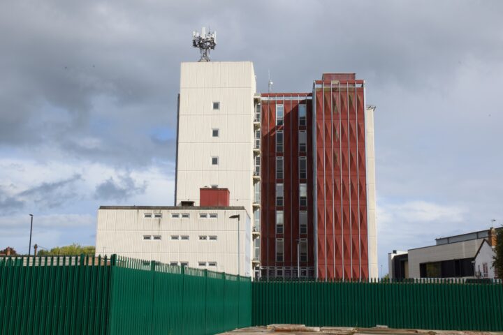 A tall, modern building with white and red vertical panels stands behind a green metal fence. The white section is taller and has aerials on the roof. The sky is cloudy, and some smaller buildings are visible on either side.