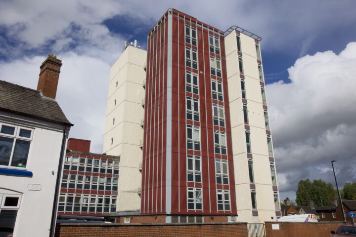 A tall, modern block of flats with vertical red and white panels stands under a partly cloudy blue sky. The building has many windows and is surrounded by a brick wall. An older, white house with a chimney is visible to the left. Trees are in the background.