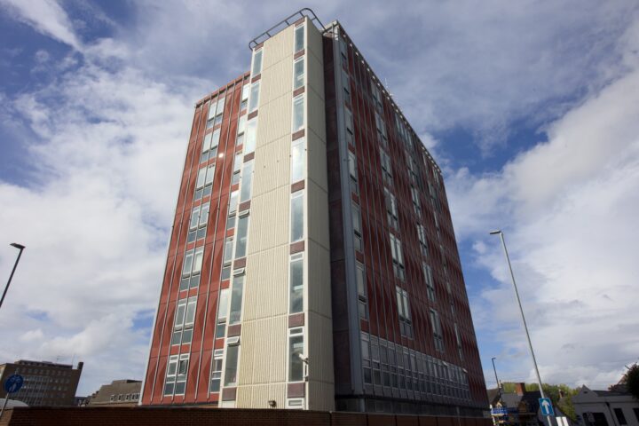 A tall, red and beige modernist block of flats with many windows is seen from below against a partly cloudy blue sky. The building stands alone, with streetlights and smaller buildings visible in the background.