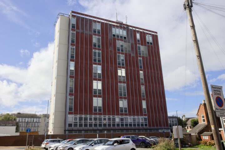 A tall, red and white office building with many windows stands under a partly cloudy sky. Several parked cars line the car park in front, and a tall wooden telegraph pole and no-parking sign are on the right side of the image. Small houses are also visible nearby.
