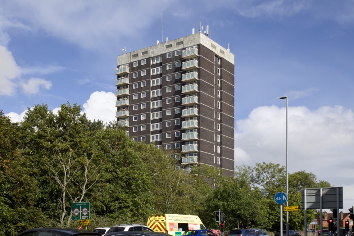 A tall, rectangular high-rise block of flats with dark brick walls and rows of balconies stands behind green trees. In front, there’s traffic, including an ambulance and several cars, under a partly cloudy blue sky. Road signs and a lamp-post are visible.