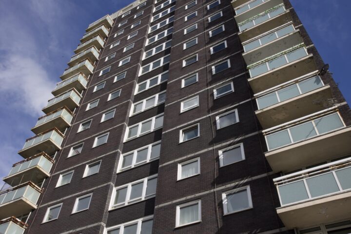 A tall, dark-brick block of flats with multiple rows of white-trimmed windows and light blue balconies is seen from a low angle against a partly cloudy blue sky. The balconies show signs of wear and peeling paint.