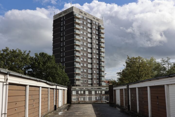 A tall, dark-brick block of flats with balconies rises behind two rows of brown and white garage doors. Trees flank the scene and clouds fill the blue sky above. The viewpoint is from the drive between the garages.