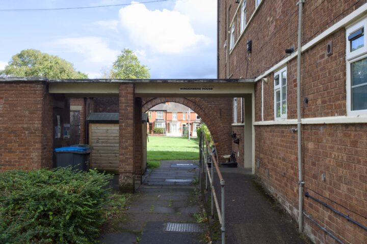 A brick archway labelled “Windermere House” connects two red-brick residential buildings. A narrow paved path and metal handrail run beneath the arch. A wooden shed and bins sit on the left. Green grass and trees are visible in the bright background.