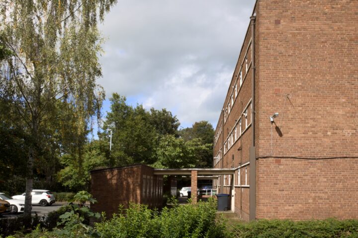 A three-storey brick building with many windows stands beside trees and bushes. A small covered walkway leads to a door, and several white cars are parked on the left. A security camera is mounted on the wall under a partly cloudy sky.