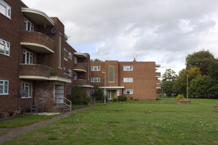Red-brick block of flats with white balconies and railings surround a large grassy courtyard. Some windows have satellite dishes. Trees and a cloudy sky are in the background, giving a suburban and tranquil atmosphere.