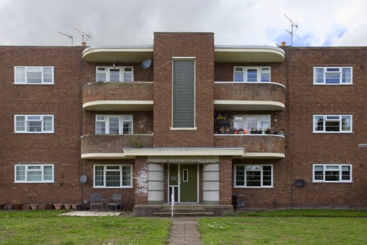 A three-storey brown brick block of flats with curved balconies, white-framed windows, and a central entrance with a glass block column. There are outdoor chairs, potted plants on balconies, and a grassy area with a path in front.