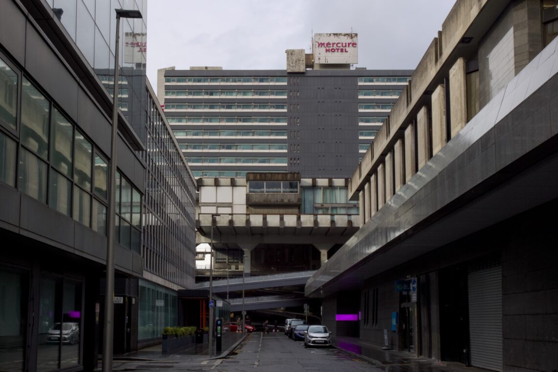 A narrow city street is flanked by modern buildings with glass and concrete façades. At the end, a tall, blocky hotel with the sign “Mercure Hotel” rises over a skybridge and flyovers, under a cloudy, grey sky. The street is empty and wet.