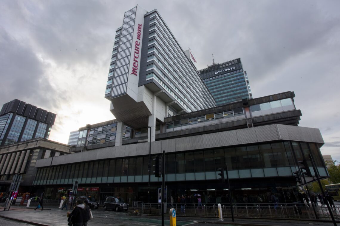 A modern, multi-storey building with Mercure Hotel in red letters on a cantilevered section. The structure has large windows and geometric shapes. Another tall building, City Tower, is visible in the background under a cloudy sky. People walk on the wet street below.