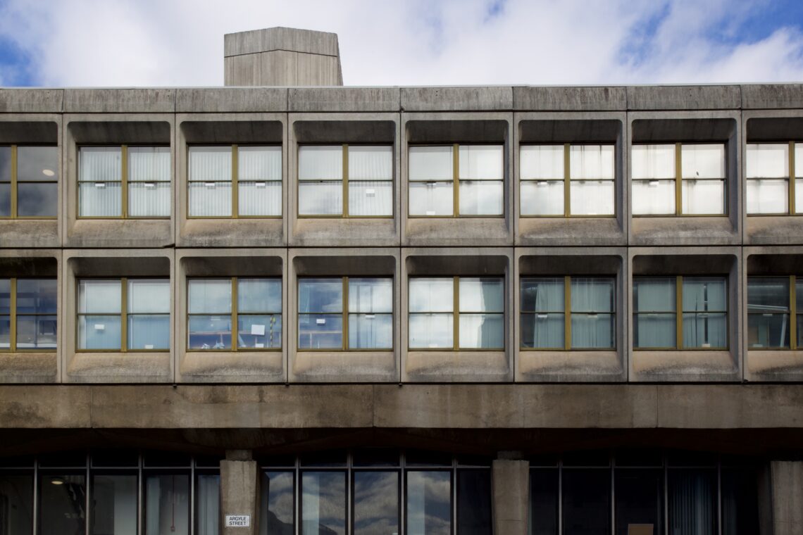 A concrete building with a Brutalist architectural style, featuring rectangular grid-like windows framed by thick concrete supports. The upper floors have evenly spaced windows, whilst the lower level has larger glass panels. Blue sky with clouds above.
