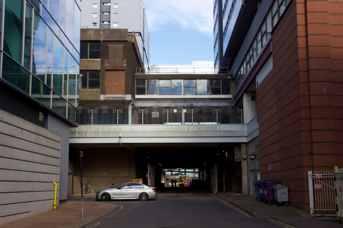 A silver car waits at a corner by a modern urban alleyway. Tall buildings with glass, brick, and concrete façades line the narrow street. Overhead, a raised walkway spans between buildings. Blue sky and clouds are visible above the scene.