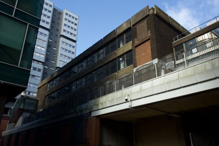 Multi-storey, modernist buildings made of concrete and brick stand under a blue sky. A tall residential tower rises in the background, while a lower building with large windows and metal fences/shutters runs alongside a raised walkway in the foreground.
