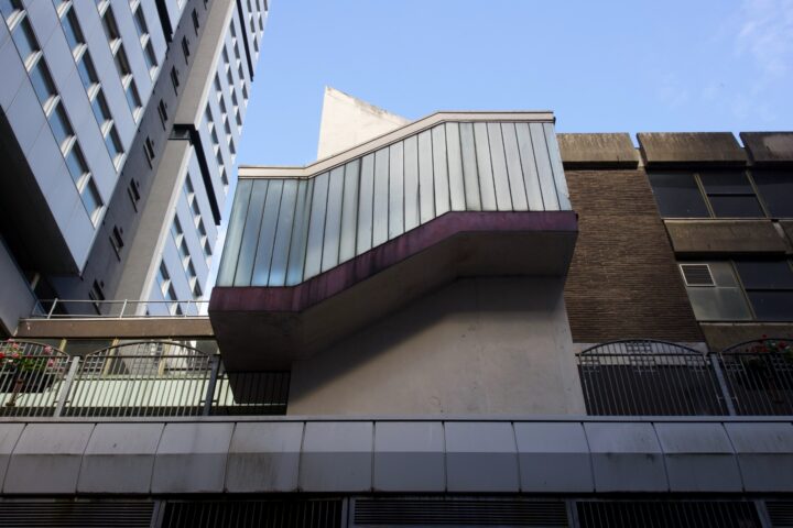 A zigzag-shaped staircase with metal cladding and glass panels juts out from a modern building facade. The structure casts a shadow on the white wall below. Surrounding buildings feature glass windows, dark bricks, and a blue sky overhead.