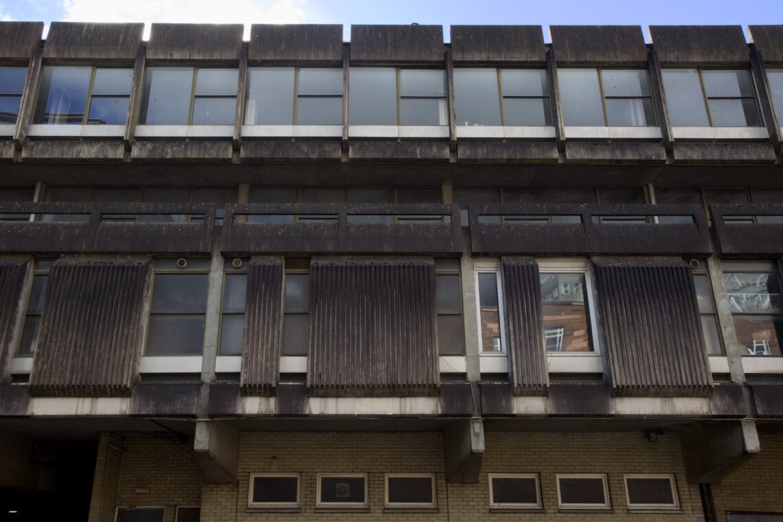 A brutalist-style concrete building with large rectangular windows, vertical ribbed panels, and weathered dark surfaces. The lower level has yellow brickwork and small square windows. Reflections of another building can be seen in some windows.