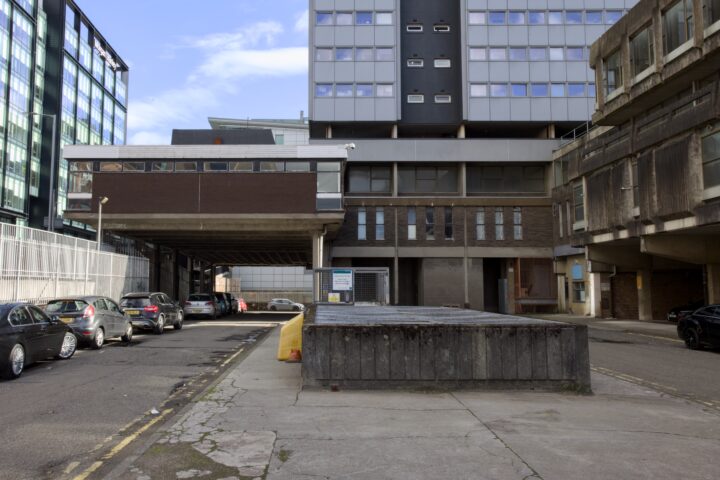 A narrow urban street flanked by modern and older concrete buildings. Several parked cars line the left side. In the centre, a low concrete structure stands on a worn pavement. The sky is mostly clear, and the overall scene appears quiet and empty.