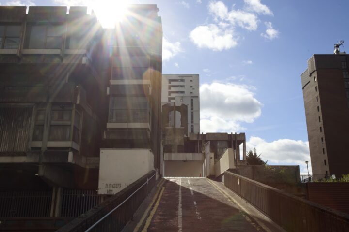 A bright sun flares over the rooftop of a brutalist concrete building. The ramp leads up between dark, weathered structures. A tall white residential block is visible in the background against a blue sky with scattered clouds.