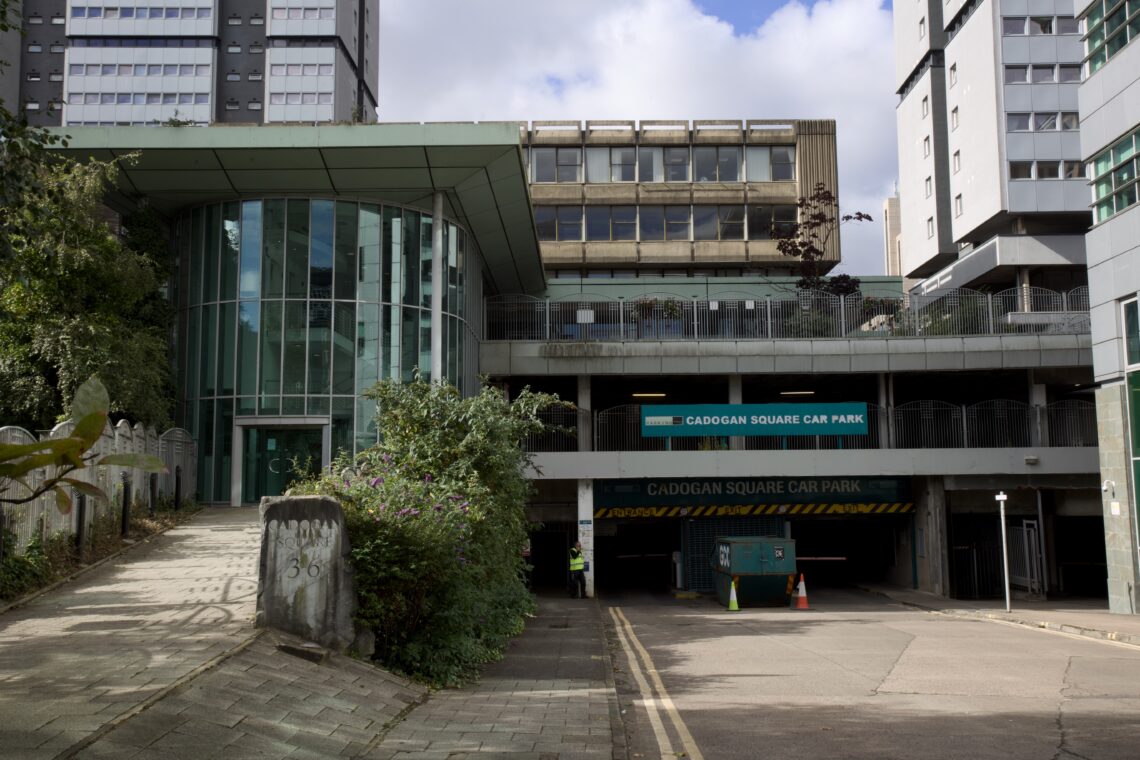 A modern multi-storey car park entrance with a sign reading Cadogan Square Car Park. Surrounding the building are tall offices, greenery, and a ramp for vehicles. Orange traffic cones and caution stripes block the entrance. The sky is partly cloudy.