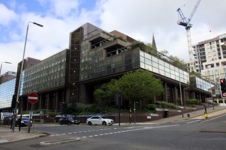 A large, modern office building with dark glass panels and greenery on upper terraces sits at a street corner. Cars are parked nearby, traffic lights and street signs are visible. A construction crane and unfinished high-rise can be seen in the background.