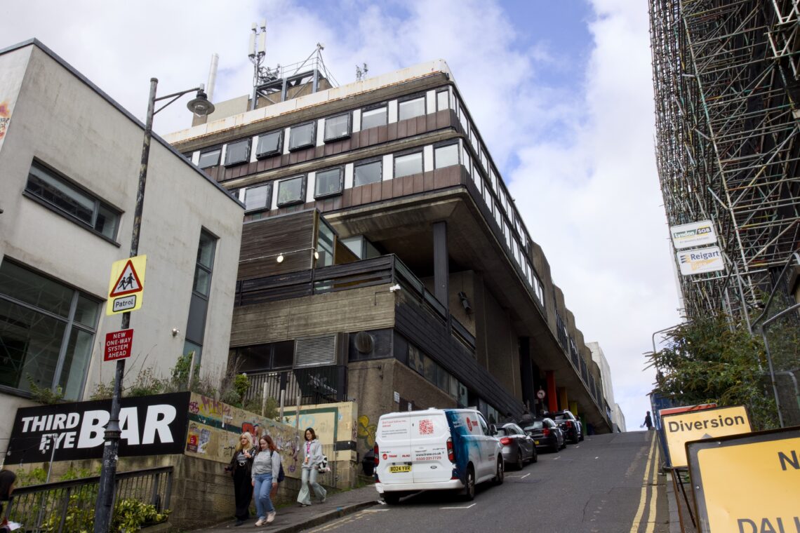 A steep street with parked cars leads uphill past a modern, concrete building. Pedestrians walk by the THIRD EYE BAR sign on the left. A yellow Diversion sign and scaffolding are on the right. The sky is partly cloudy.