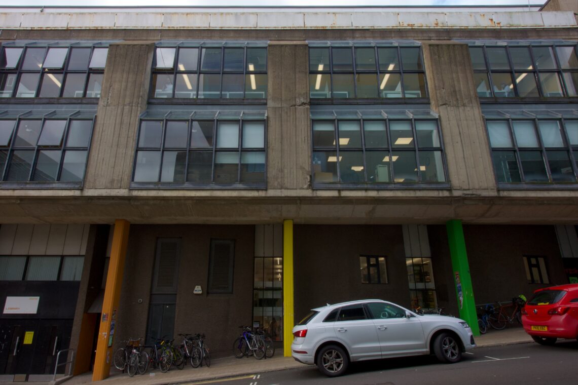 A modern building with large windows and concrete columns, some painted yellow and green. Bicycles are parked against the wall, and a white SUV and a red car are parked on the street in front. The sky is overcast.