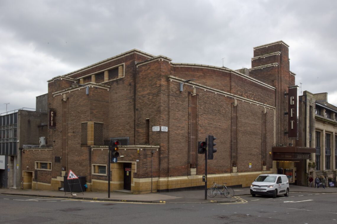 A large, brown brick building with art deco features on a street corner. The marquee reads “GFT” in vertical letters. There are traffic lights, a white van, bicycles, and a few people near the entrance. The sky is overcast.