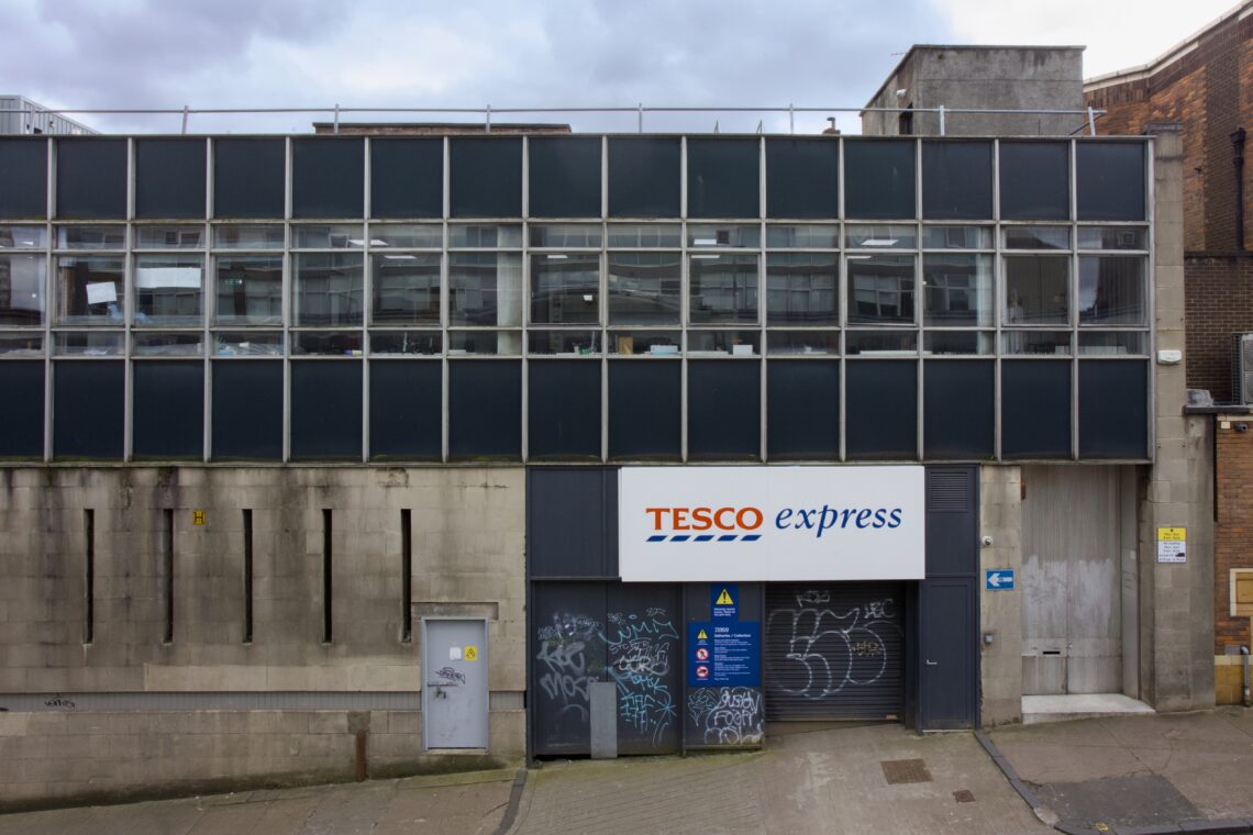 A Tesco Express shopfront with a white and blue sign is set in a grey, industrial building with large glass windows. Graffiti covers the doors and walls. The pavement in front is empty and the sky above is cloudy.