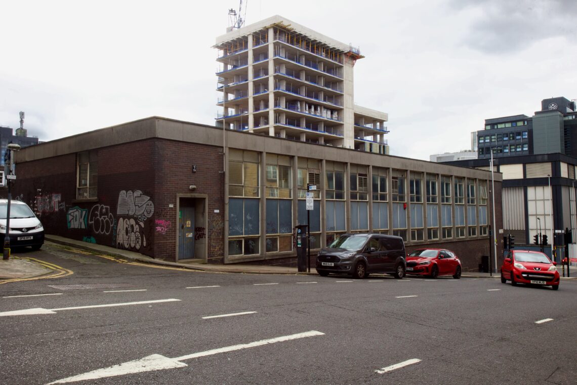 A sloped city street with parked cars beside a low, brick building covered in graffiti. Behind it, a tall building is under construction, showing exposed floors. The sky is cloudy, and several modern buildings appear in the background.