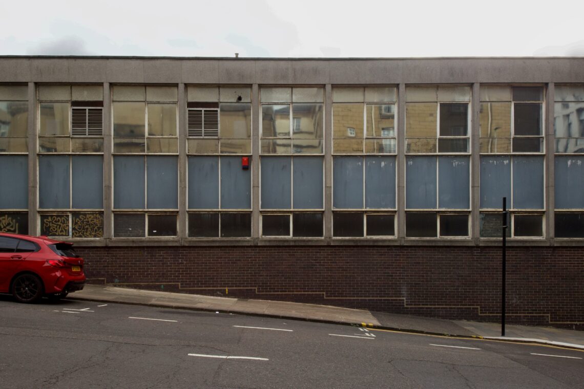 A dull, rectangular, concrete building with large, evenly spaced windows, many of which are grimy or covered. A red car is parked on a sloping street in front, and graffiti is visible on the lower windowpanes. The scene appears urban and bleak.