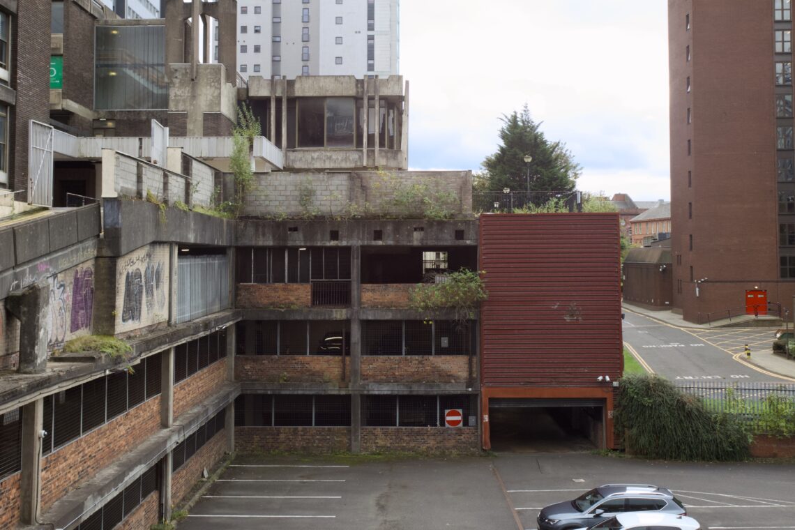 A partially abandoned, multi-storey car park with exposed brickwork, overgrown weeds, and graffiti. The car park is next to modern buildings, with a few cars parked below and a “no entry” sign at the entrance. The scene is quiet and urban.