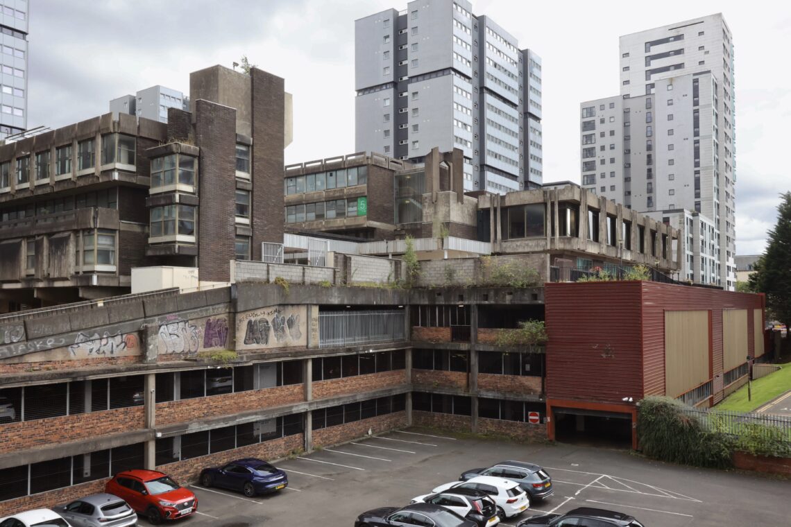 A multi-storey, concrete car park with graffiti on its walls sits alongside modern high-rise buildings. Several parked cars fill the car park. The older, weathered structure contrasts sharply with the new, sleek residential towers in the background.